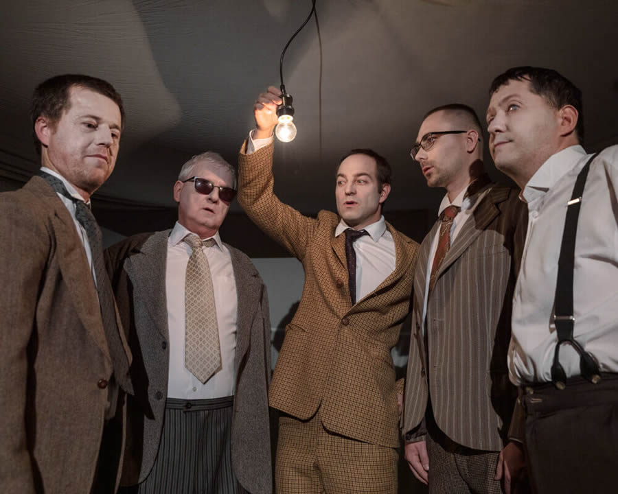 Päť hercov v polkruhu smeruje svoj pohľad na žiarovku uprostred nich